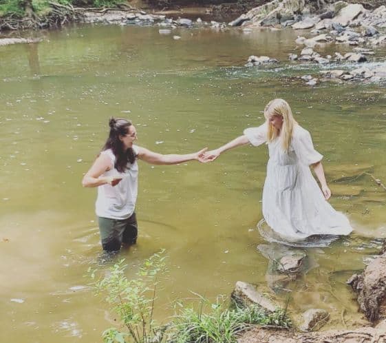 Elizabeth in white dress wades into the river, with Melissa standing in the river holding her outstretched hand.