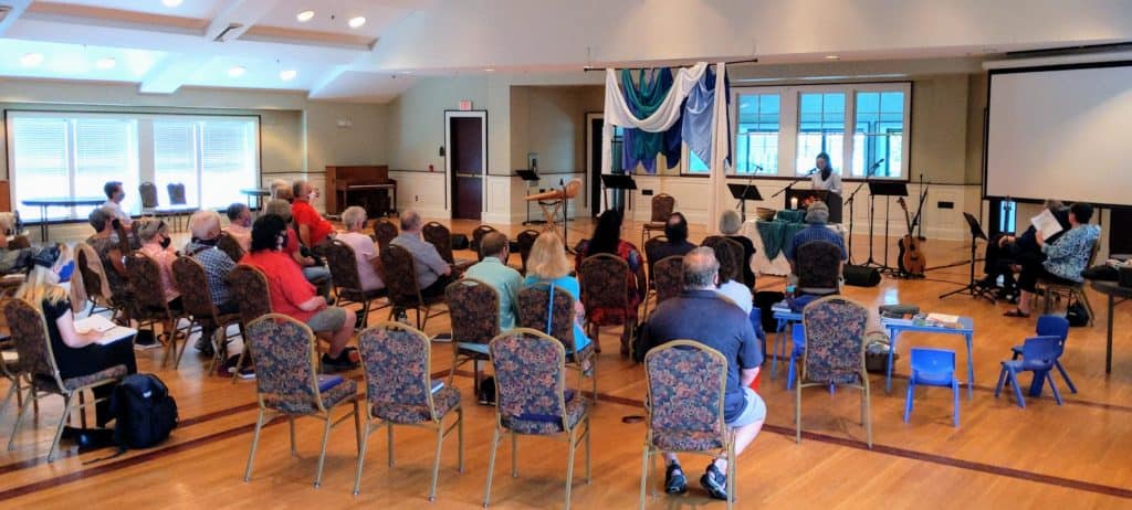 congregation sitting in chairs facing Melissa who is preaching at the front of the hall.