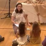 Melissa sitting on the floor telling a story to children who have their hands raised.