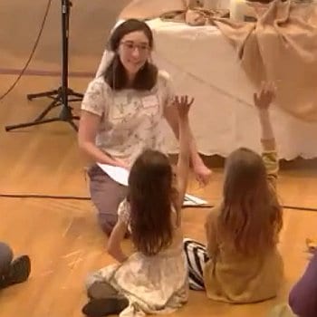 Melissa sitting on the floor telling a story to children who have their hands raised.