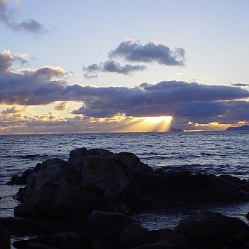 A dark ocean scape with craggy rocks in the foreground, clouds in the sky with the sun's rays shining out from behind a dark cloud.