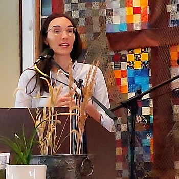 Melissa preaching at RMC on March 19, 2023, with a quilt behind her and a plant and stalks of brown grass and wheat on a table in front of her.