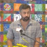 Raleigh Mennonite Church Chas Edens, a man with dark hair and a beard preaches at Raleigh Mennonite in front of a colorful banner.