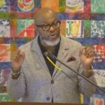 Raleigh Mennonite Church Dr Derek Hicks preaching at Raleigh Mennonite in front of a colorful quilted wall hanging, July 30, 2023.