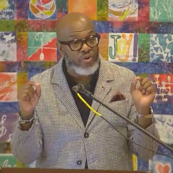 Dr Derek Hicks preaching at Raleigh Mennonite in front of a colorful quilted wall hanging, July 30, 2023.