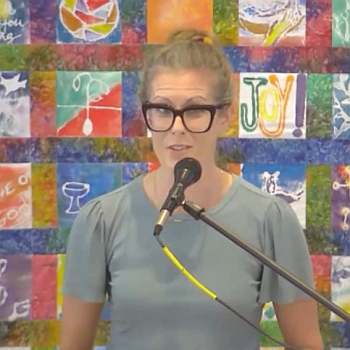 Elizabeth Korver-Glenn speaking at Raleigh Mennonite in front of a colorful quilted wall hanging on Aug. 16, 2023