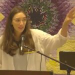 Raleigh Mennonite Church Melissa preaching with her hand raised outward. A star-patterned quilt is behind her.