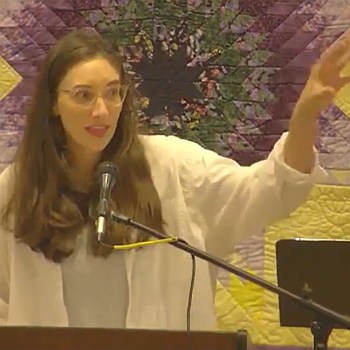 Melissa preaching with her hand raised outward. A star-patterned quilt is behind her.