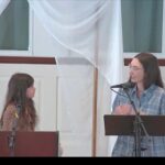 Melissa and Juliet preaching at Raleigh Mennonite. Melissa standing behind a music stand, is looking at Juliet who is behind a podium.