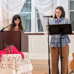 Juliet standing behind the podium and Melissa standing in front of a music stand. 