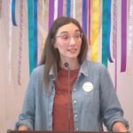 Raleigh Mennonite Church Melissa preaching in front of a banner of ribbons on June 16, 2024