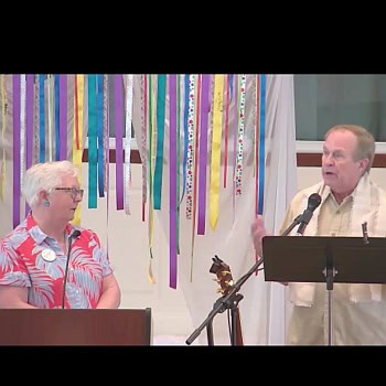 Susan listening to Hans speak at RMC on June 30, 2024. They're in front of a banner with ribbons.