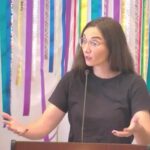 Raleigh Mennonite Church Melissa, holding her hands out, preaching on Aug. 11, 2024 in front of a banner of colorful ribbons.