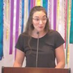 Raleigh Mennonite Church Melissa preaching in front of a banner of ribbons on Aug. 18, 2024