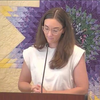 Melissa, in a white shirt, preaching in front of a quilt wall hanging.