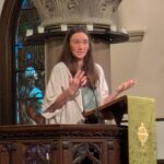 Raleigh Mennonite Church Melissa preaching in the sanctuary at Church of the Good Shepherd. She stands in the podium with her hands in front of her with a marble pillar seen in the background.