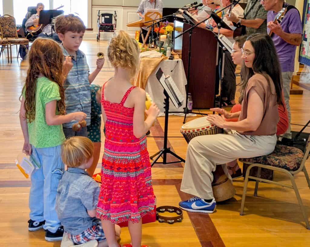 Melissa plays djembe while children play rhythm instruments in front of her.