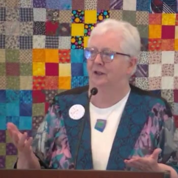Susan speaking on Sunday, Sept. 21, 2025 in front of a colorful quilted back drop.