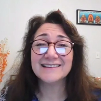 Woman with long hair and glasses speaking on zoom with a small picture on the wall behind her