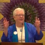 Susan wearing a blue jacket, holding up "air quotes" while preaching in front of a star-shaped quilted wall hanging.