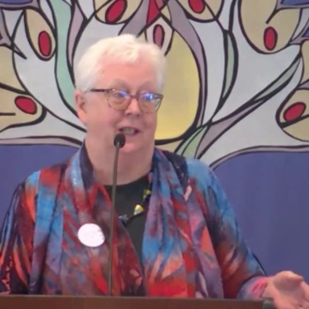 Susan preaching at RMC on May 3, 2026 in front of a banner with a sprouting flower on it.