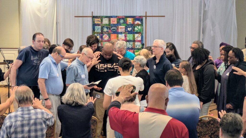 Many people gather around Trey with heads bowed, laying their hands on him as Doug Luginbill provides a prayer of dedication for Trey's ministry 