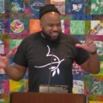 Trey preaching with hands in motion while wearing a black beanie and a black t-shirt with a white dove on it.