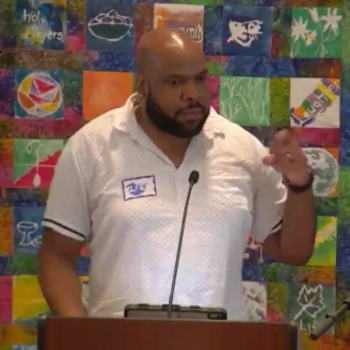 Trey speaking with a hand raised for emphasis at RMC in front of a colorful quilt.